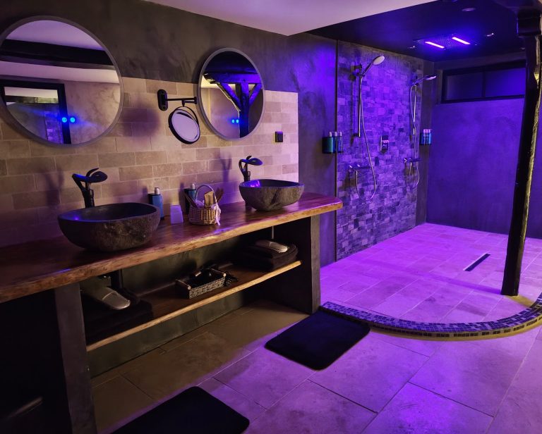 Salle de bain XXL, pierre et bois nature Salle de bains moderne avec deux lavabos plateau en chêne naturel, douches, ciel de pluie et éclairage violet. sol en travertin